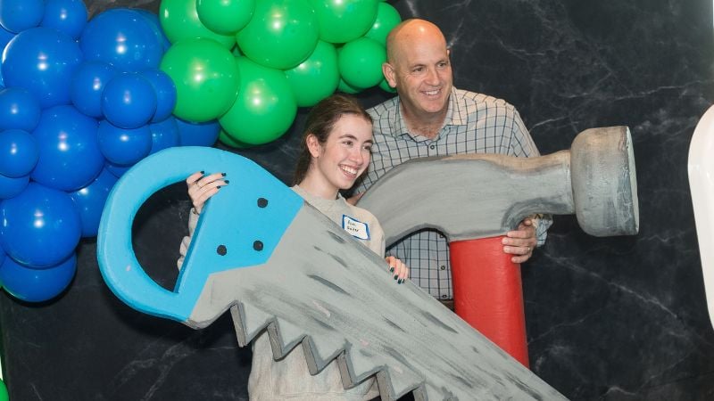 Father and daughter posing with prop hammer and saw in a photo booth with bright baloons.