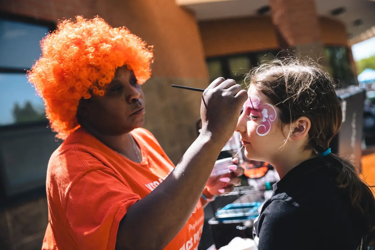 A woman in a bright orange wig and matching t-shirt, painting a child's face with pink and white face paint.